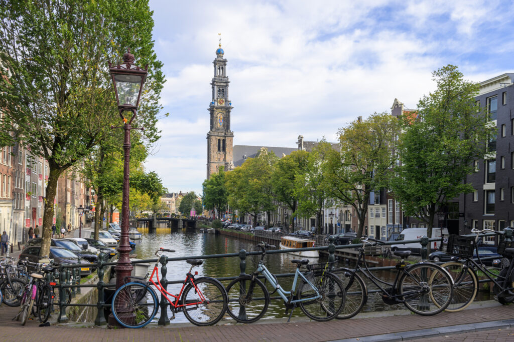 Sfeervolle stadsfotografie van de Prinsengracht in Amsterdam met geparkeerde fietsen op de brug en de iconische Westerkerk onder een blauwe lucht.