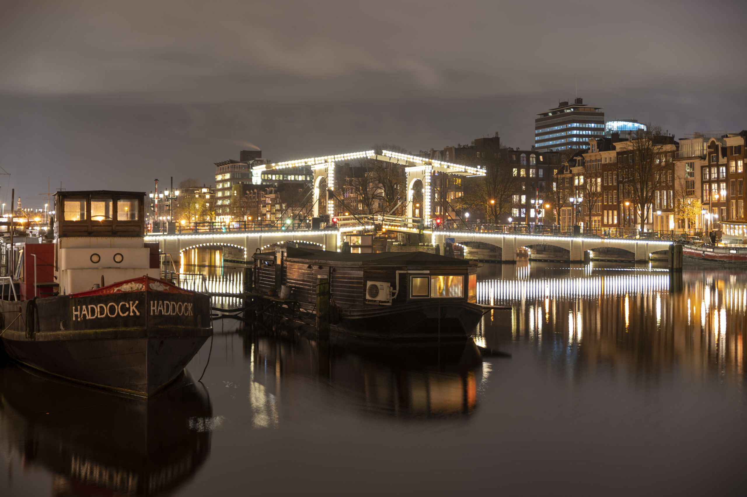 Sfeervolle nachtfotografie als exclusief schilderij van de verlichte Magere Brug over de Amstel in Amsterdam met woonboten en reflecties op het water. Verkrijgbaar als luxe wanddecoratie bij FotoAmsterdam